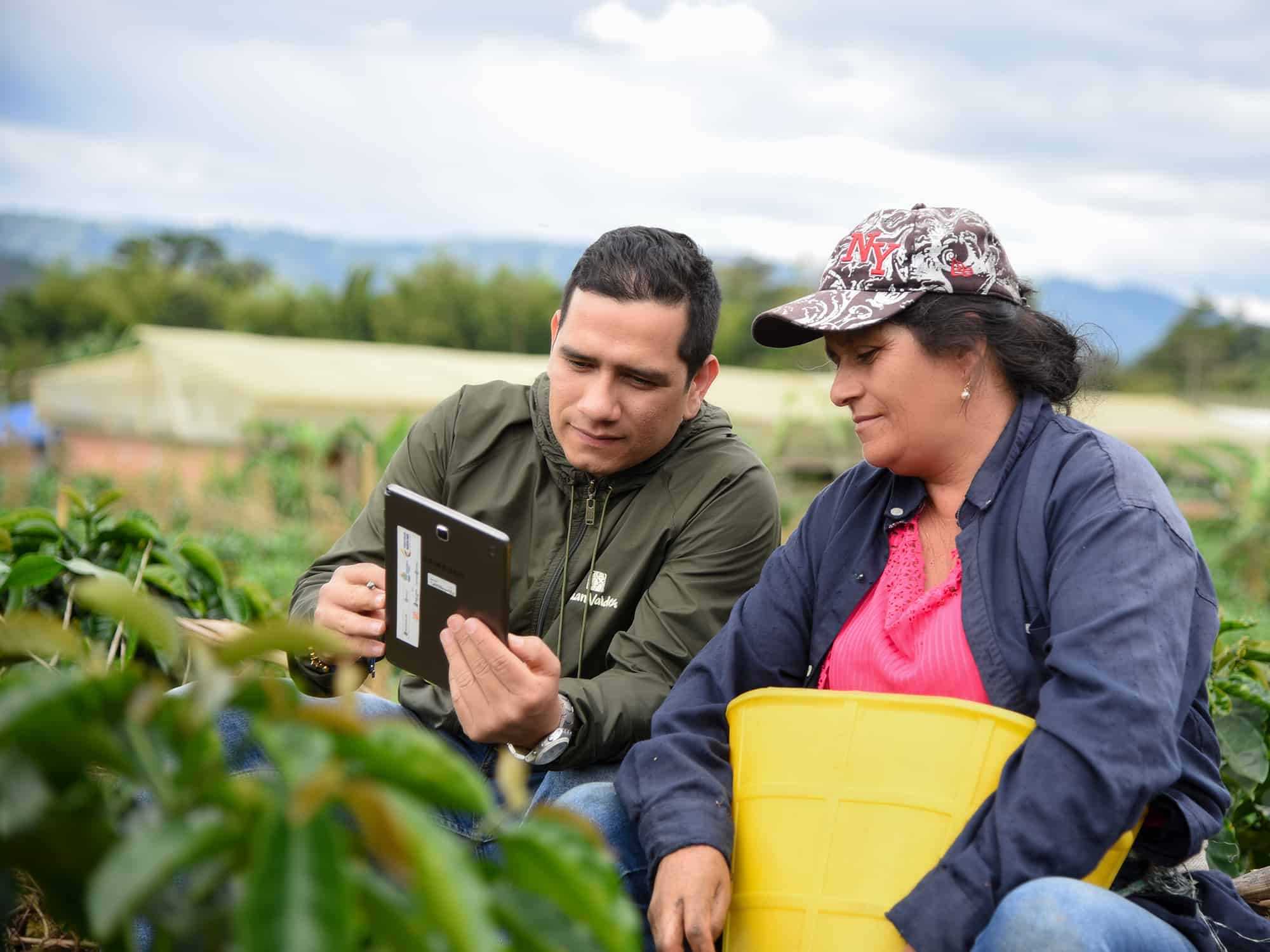 A man holding a tablet and a woman looking at the tablet, both are sitting in a green field